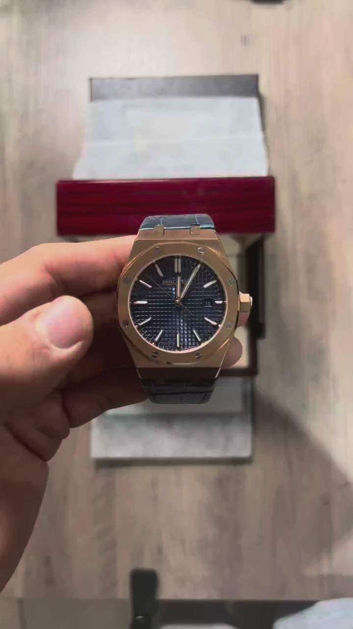 Reloj octagonal con caja en tono oro rosa, esfera azul con textura de cuadrícula y correa de piel azul, sostenido en la mano frente a una caja de presentación roja