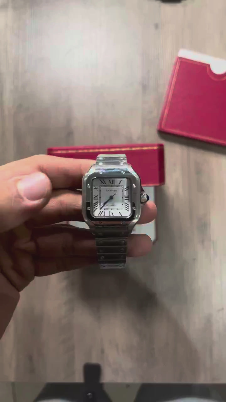 Reloj cuadrado de acero con esfera blanca, números romanos y manecillas azules, sostenido en la mano frente a una caja roja