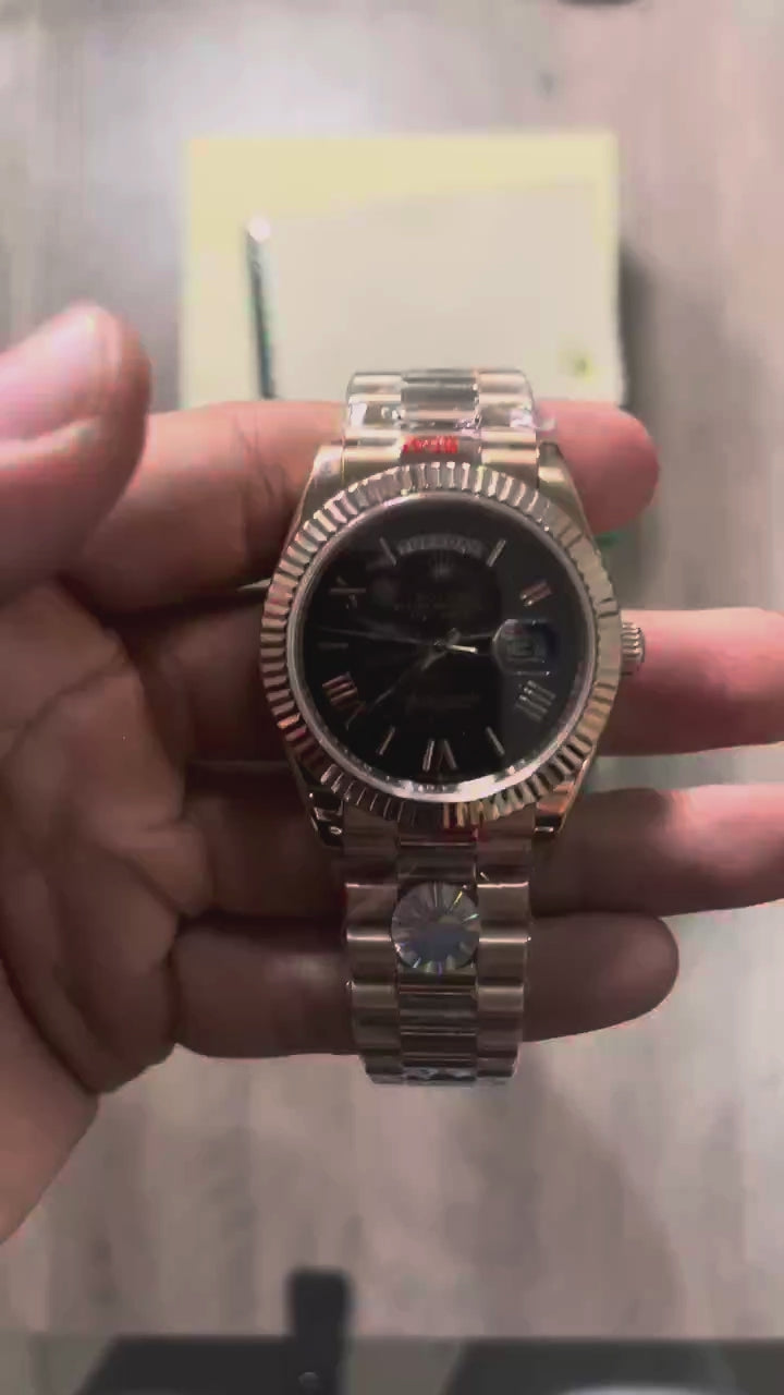 Reloj en tono oro rosa con esfera negra, bisel estriado y brazalete de eslabones, sostenido en la mano frente a una caja verde