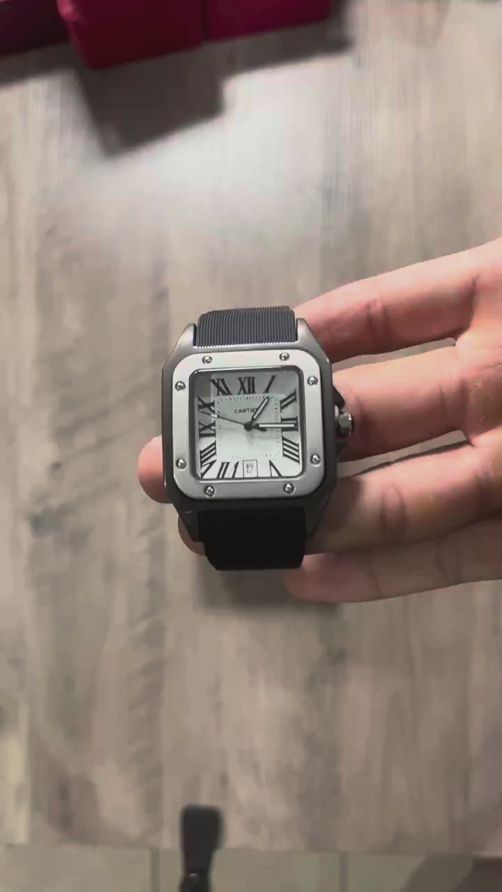 Reloj cuadrado de caja negra con bisel plateado, esfera plateada con números romanos y correa de caucho negro, sostenido en la mano