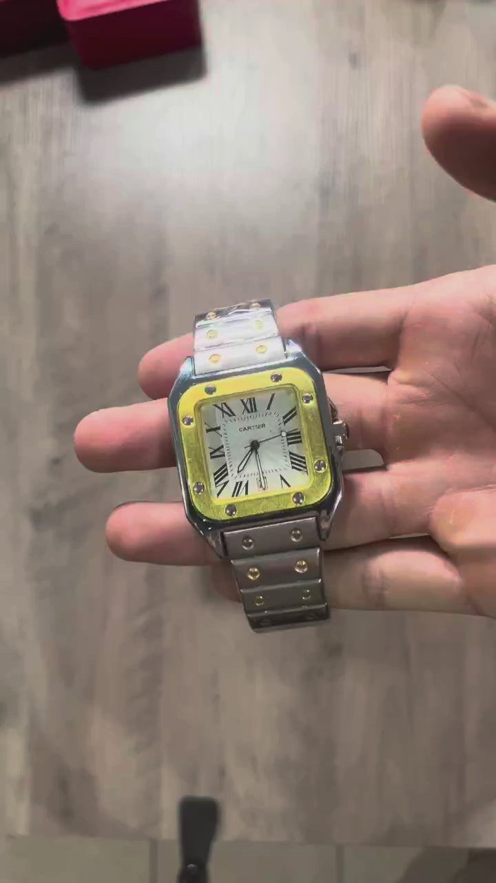 Reloj cuadrado de acero con bisel dorado, esfera clara con números romanos y brazalete con tornillos dorados, junto a una caja roja sobre fondo de madera