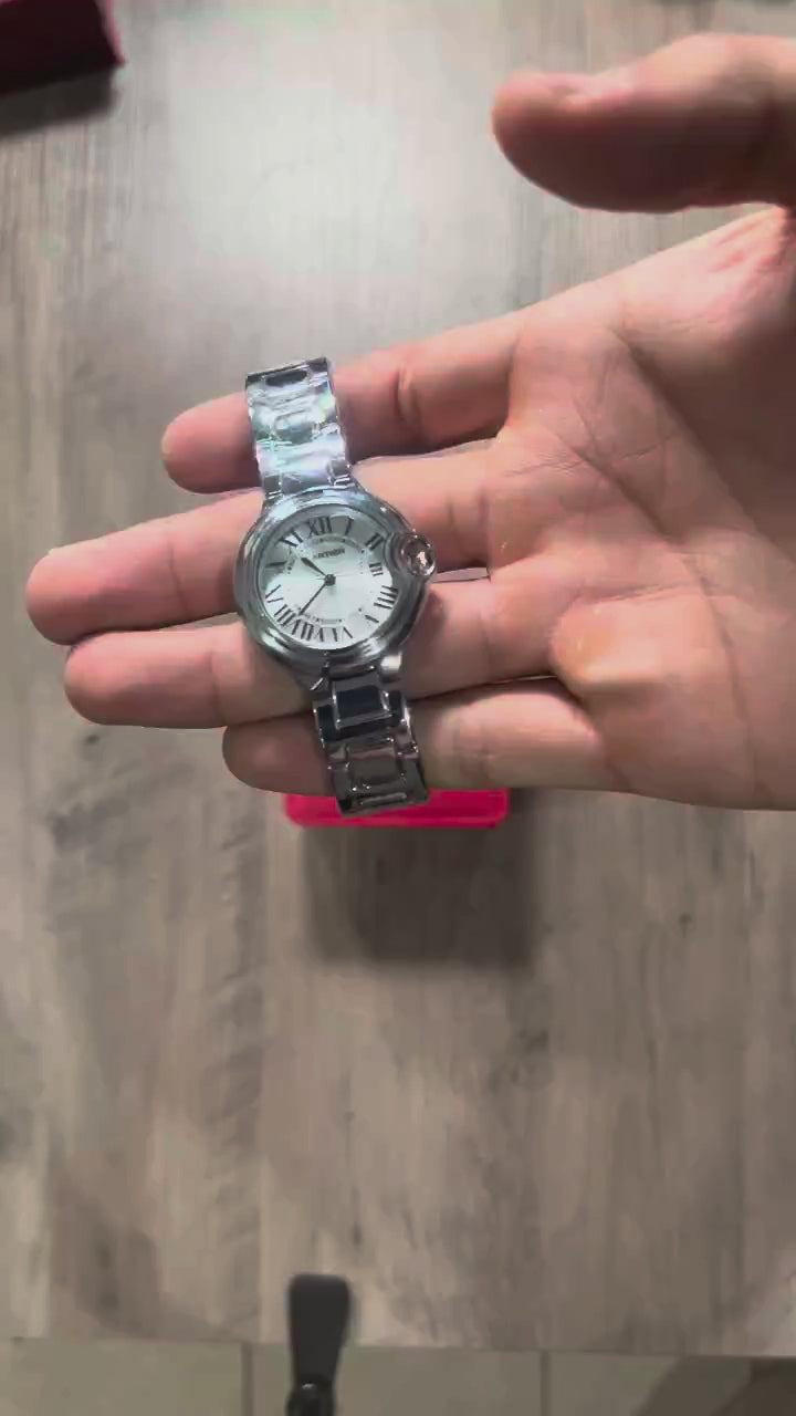 Reloj redondo plateado de acero con esfera plateada, números romanos y brazalete de eslabones, sostenido en la mano