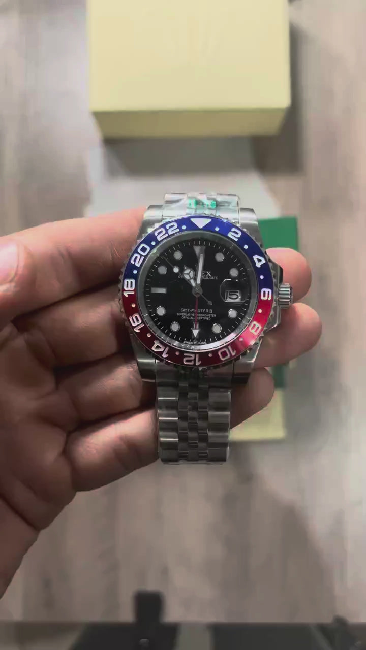 Reloj de acero con bisel azul y rojo, esfera negra con fecha y brazalete de cinco eslabones, sostenido en la mano