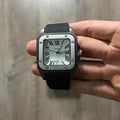 Reloj cuadrado de caja negra con bisel plateado, esfera plateada con números romanos y correa de caucho negro, sostenido en la mano