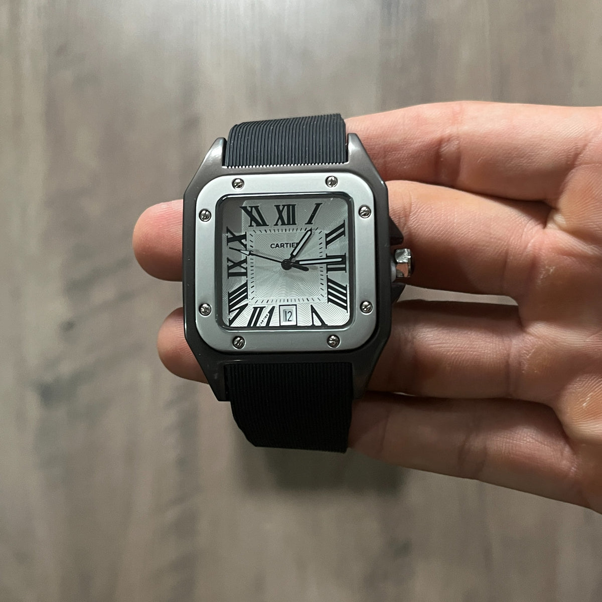 Reloj cuadrado de caja negra con bisel plateado, esfera plateada con números romanos y correa de caucho negro, sostenido en la mano