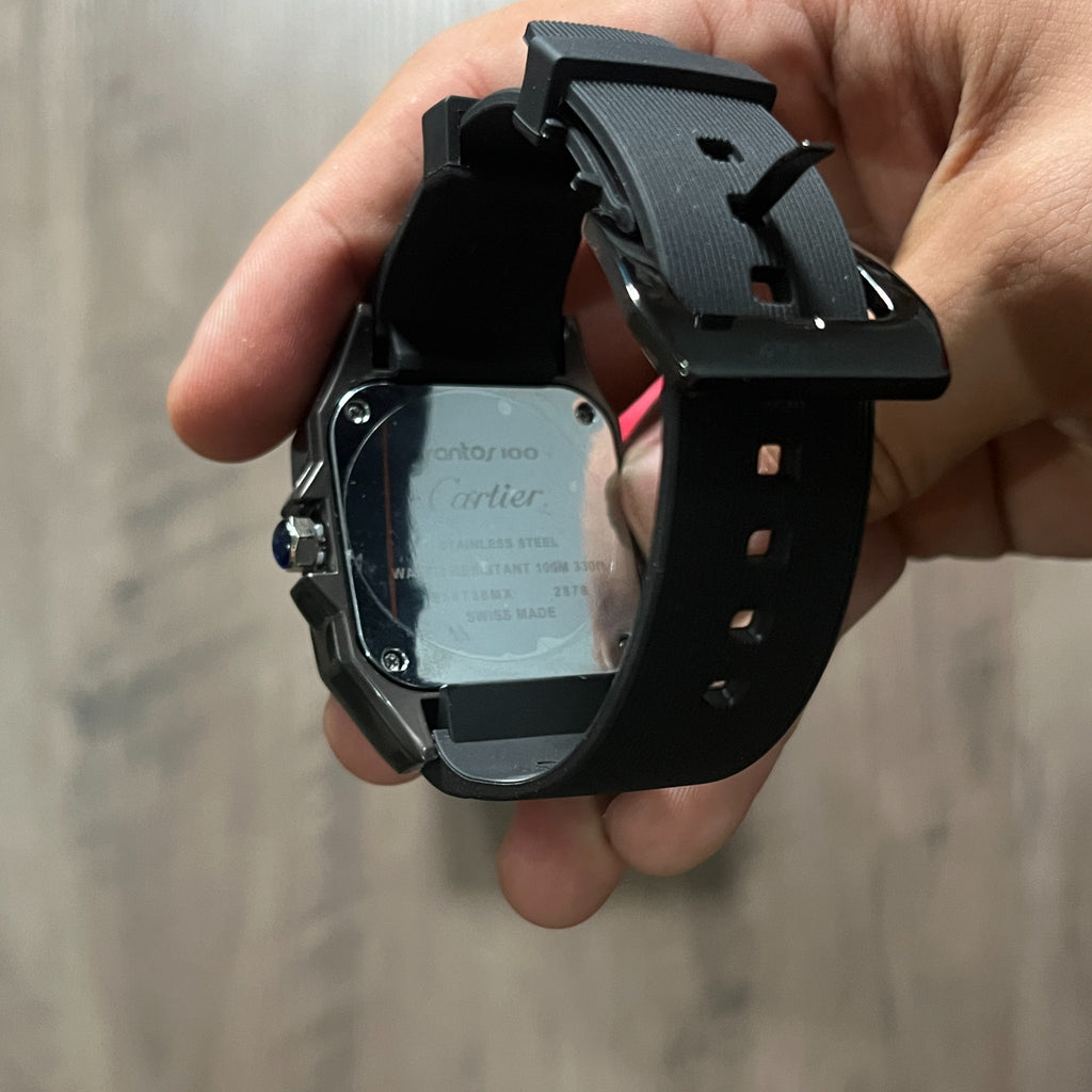 Reloj cuadrado de caja negra con bisel plateado, esfera plateada con números romanos y correa de caucho negro, sostenido en la mano