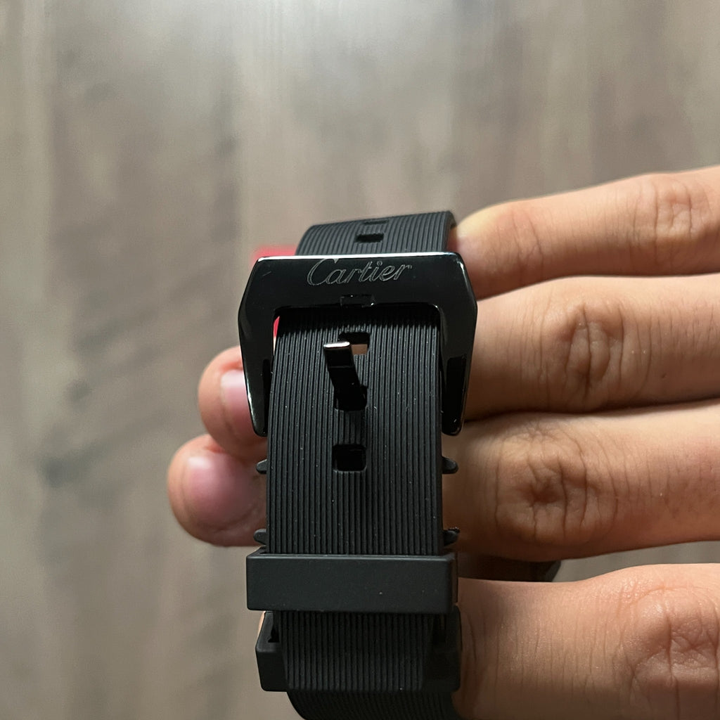 Reloj cuadrado de caja negra con bisel plateado, esfera plateada con números romanos y correa de caucho negro, sostenido en la mano