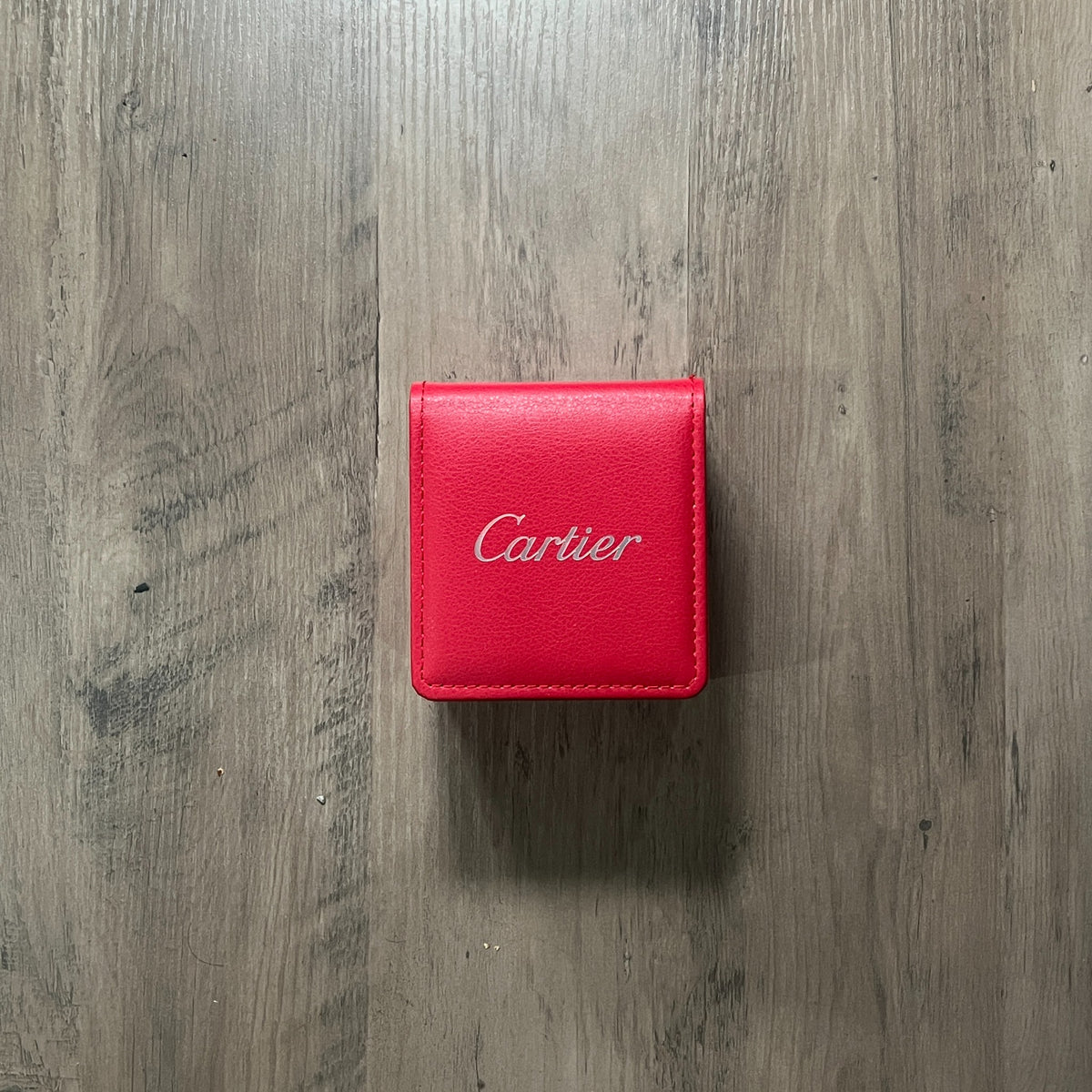 Reloj cuadrado de acero con bisel dorado, esfera clara con números romanos y brazalete con tornillos dorados, junto a una caja roja sobre fondo de madera