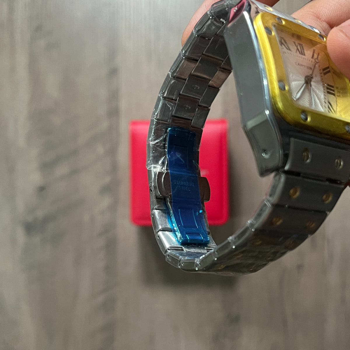 Reloj cuadrado de acero con bisel dorado, esfera clara con números romanos y brazalete con tornillos dorados, junto a una caja roja sobre fondo de madera