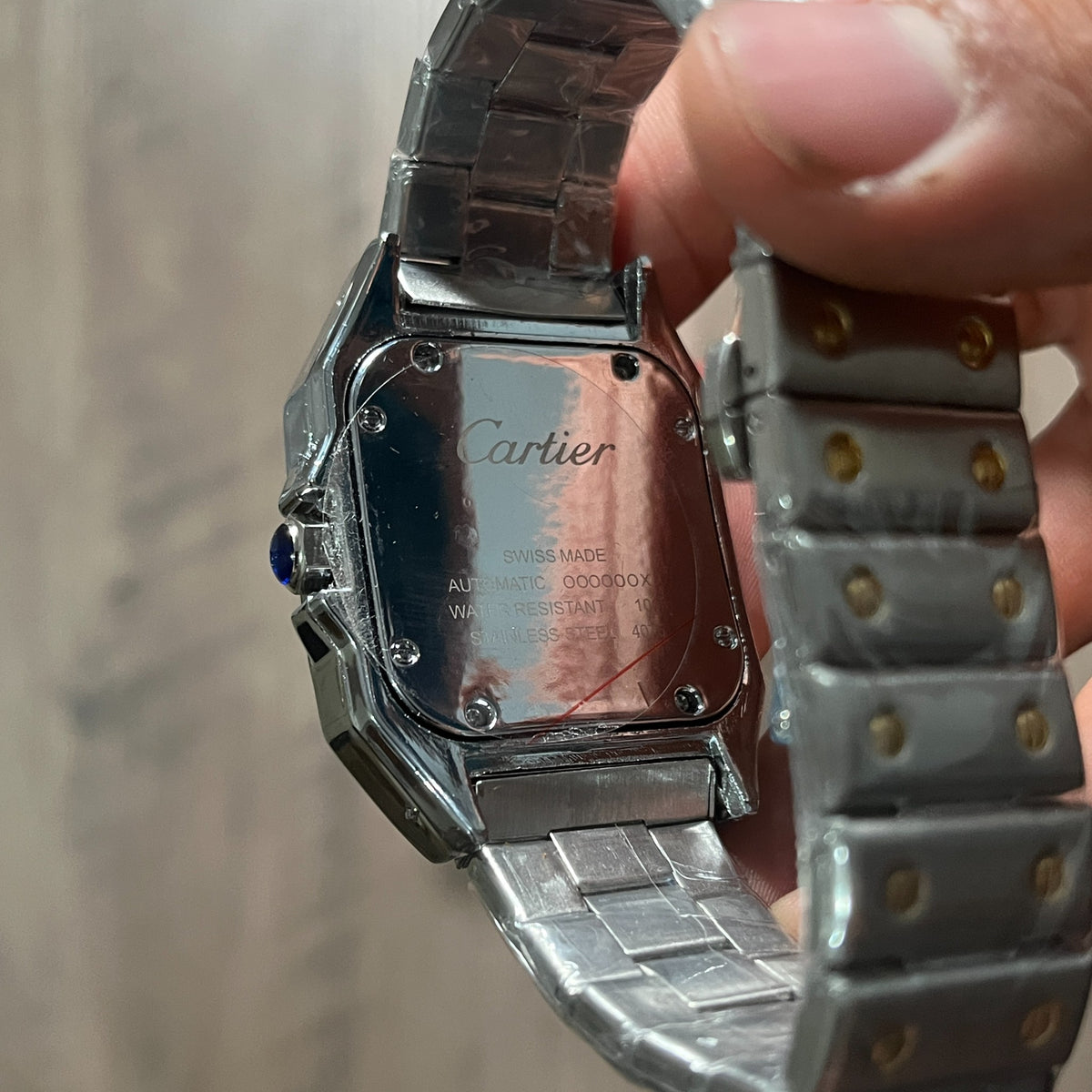 Reloj cuadrado de acero con bisel dorado, esfera clara con números romanos y brazalete con tornillos dorados, junto a una caja roja sobre fondo de madera