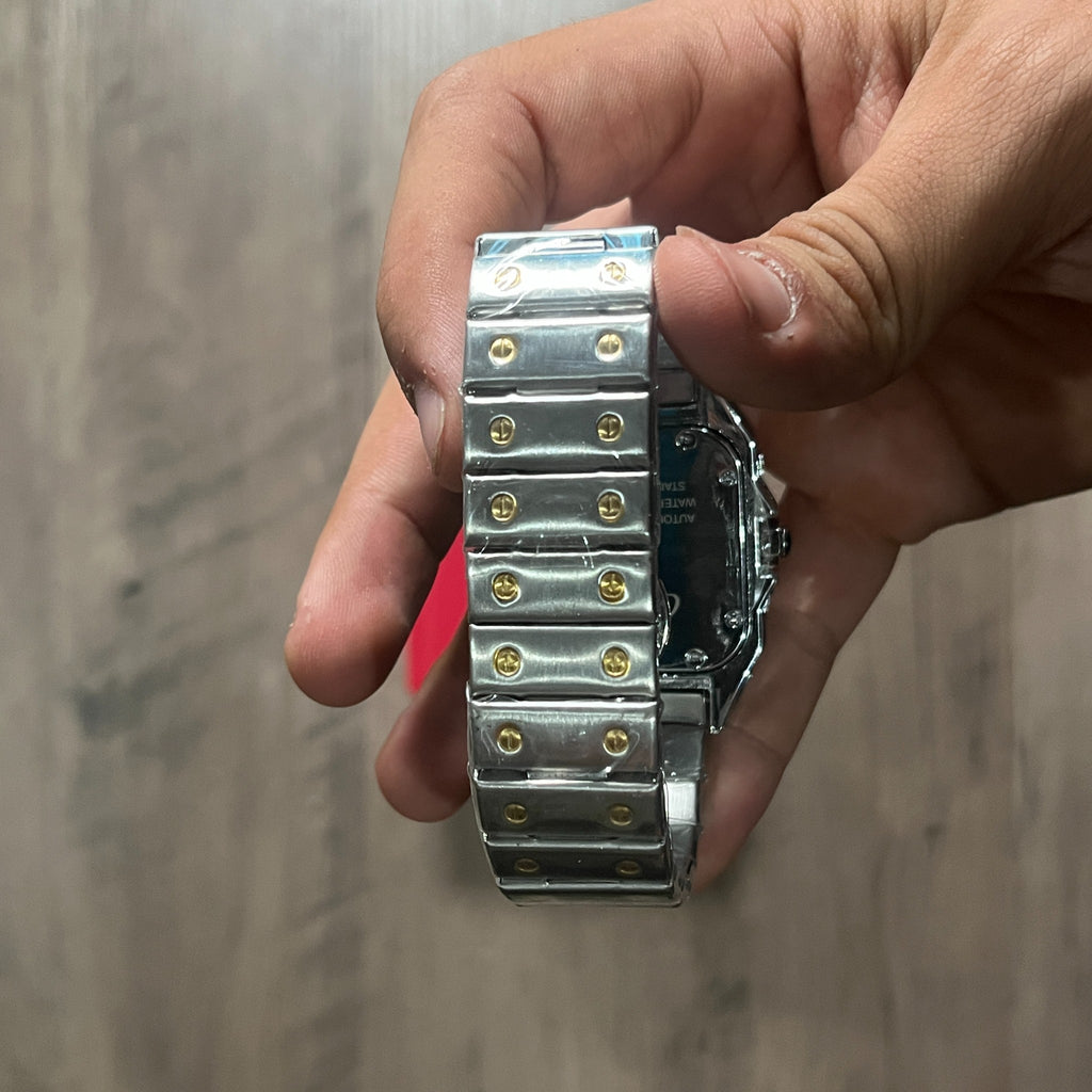 Reloj cuadrado de acero con bisel dorado, esfera clara con números romanos y brazalete con tornillos dorados, junto a una caja roja sobre fondo de madera