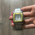 Reloj cuadrado de acero con bisel dorado, esfera clara con números romanos y brazalete con tornillos dorados, junto a una caja roja sobre fondo de madera