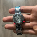 Reloj redondo plateado de acero con esfera plateada, números romanos y brazalete de eslabones, sostenido en la mano