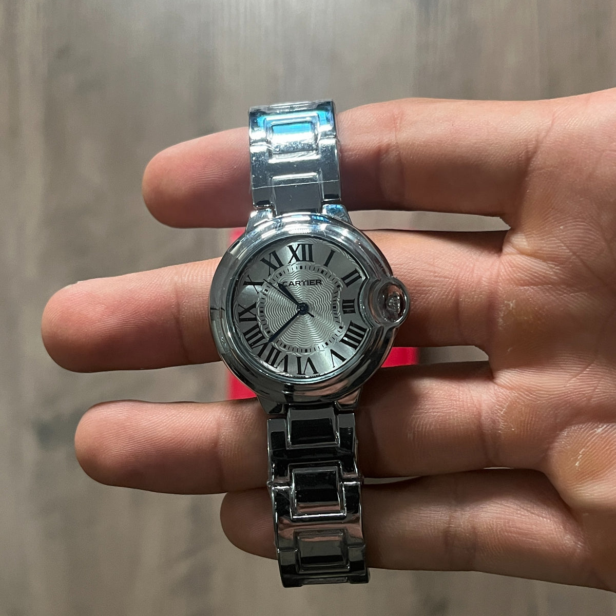 Reloj redondo plateado de acero con esfera plateada, números romanos y brazalete de eslabones, sostenido en la mano