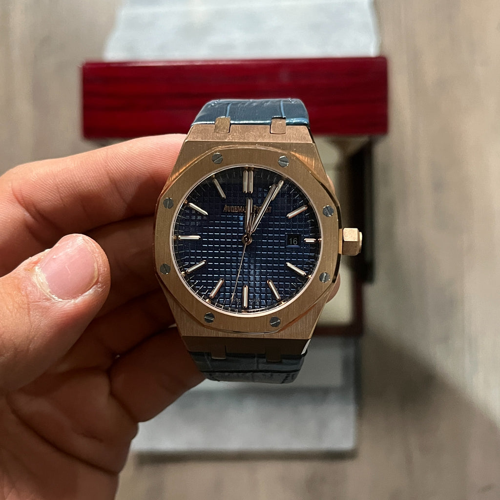 Reloj octagonal con caja en tono oro rosa, esfera azul con textura de cuadrícula y correa de piel azul, sostenido en la mano frente a una caja de presentación roja