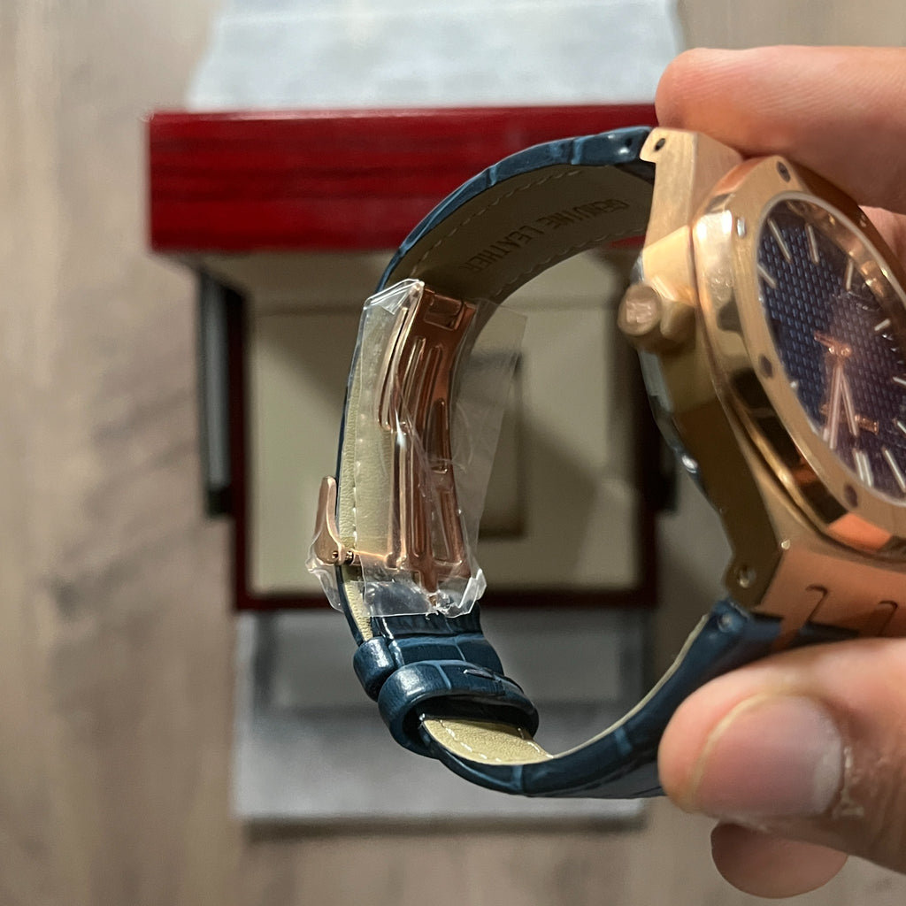 Reloj octagonal con caja en tono oro rosa, esfera azul con textura de cuadrícula y correa de piel azul, sostenido en la mano frente a una caja de presentación roja