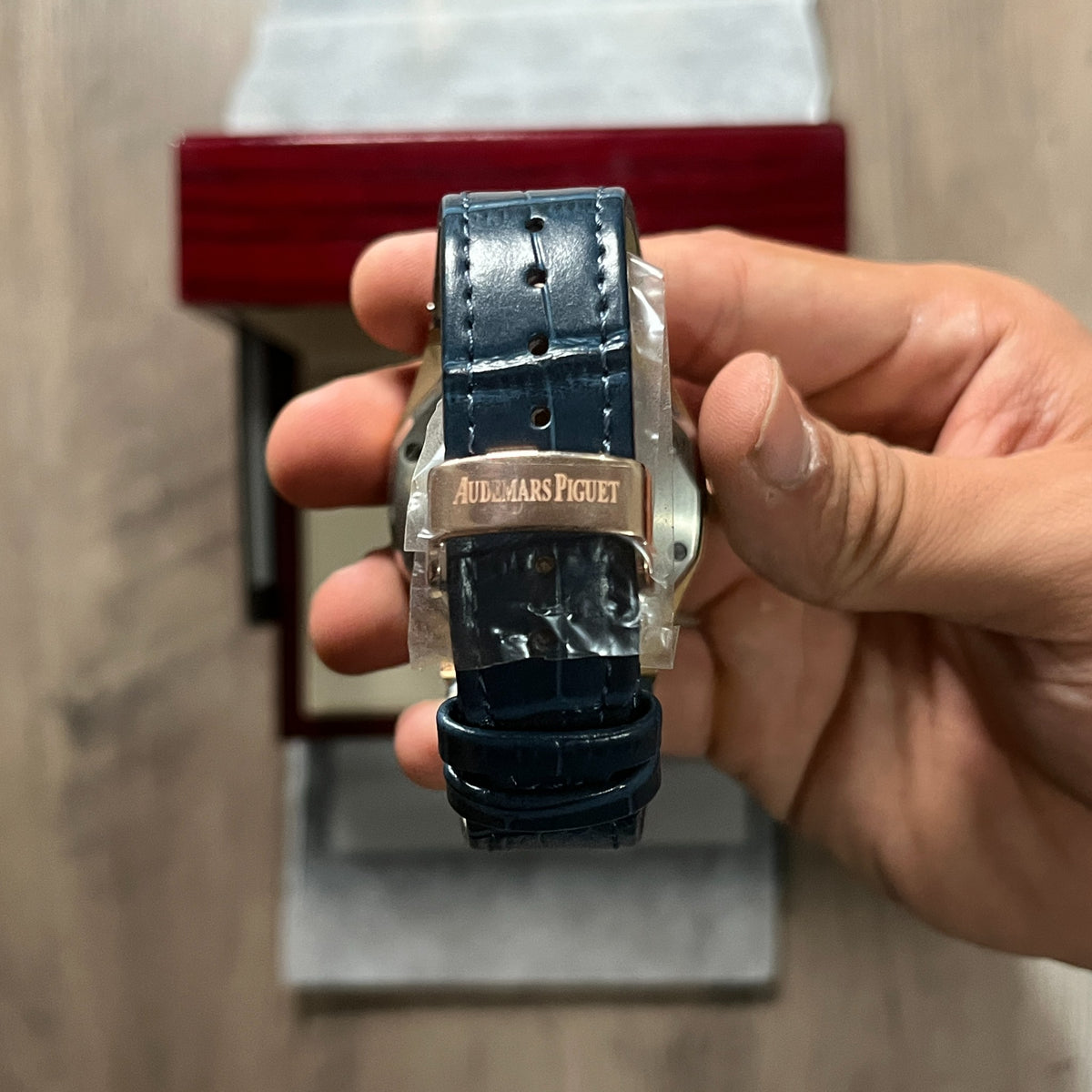 Reloj octagonal con caja en tono oro rosa, esfera azul con textura de cuadrícula y correa de piel azul, sostenido en la mano frente a una caja de presentación roja