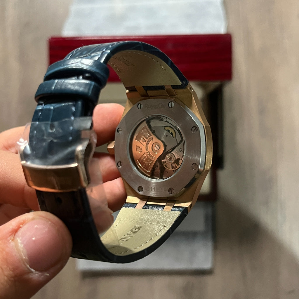 Reloj octagonal con caja en tono oro rosa, esfera azul con textura de cuadrícula y correa de piel azul, sostenido en la mano frente a una caja de presentación roja
