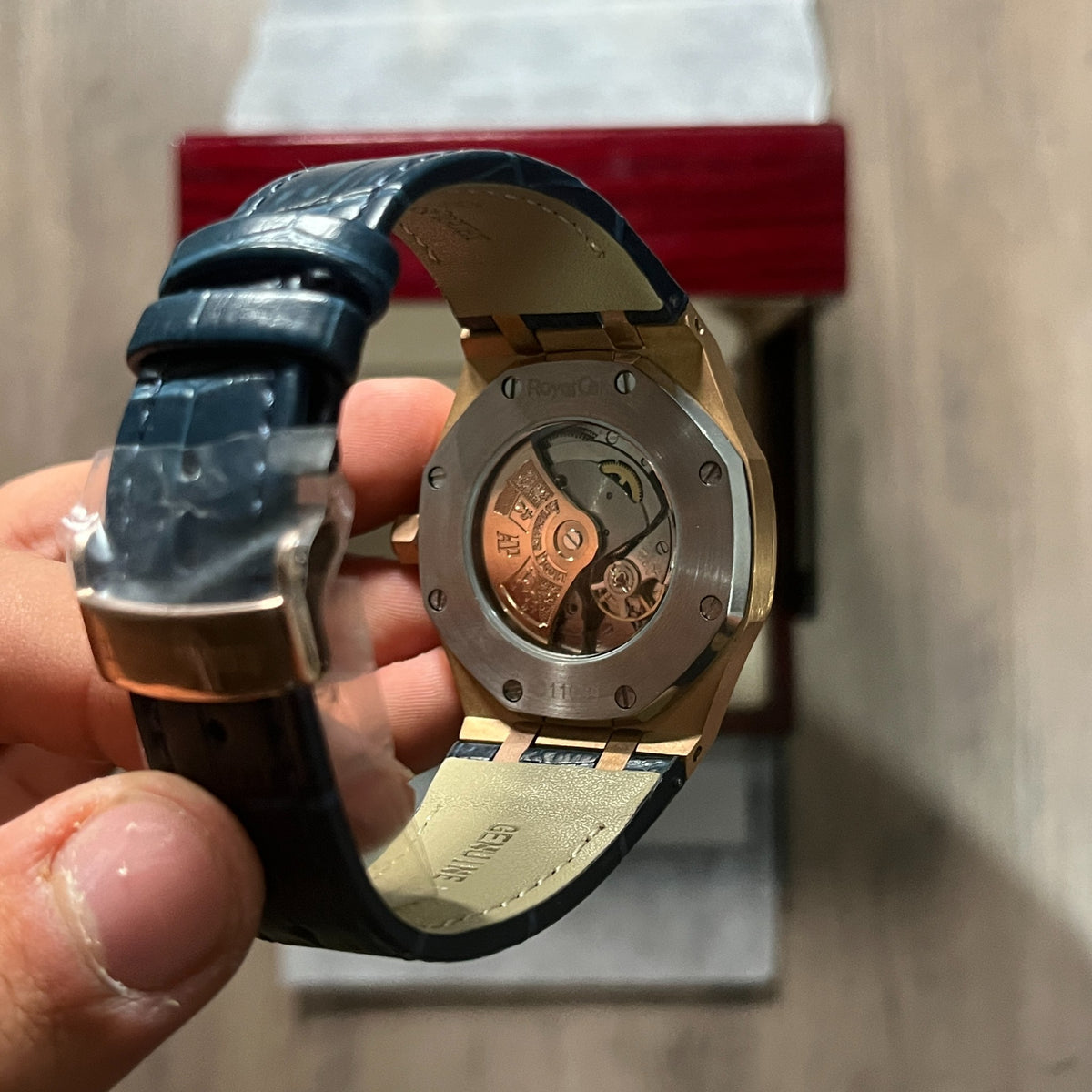 Reloj octagonal con caja en tono oro rosa, esfera azul con textura de cuadrícula y correa de piel azul, sostenido en la mano frente a una caja de presentación roja