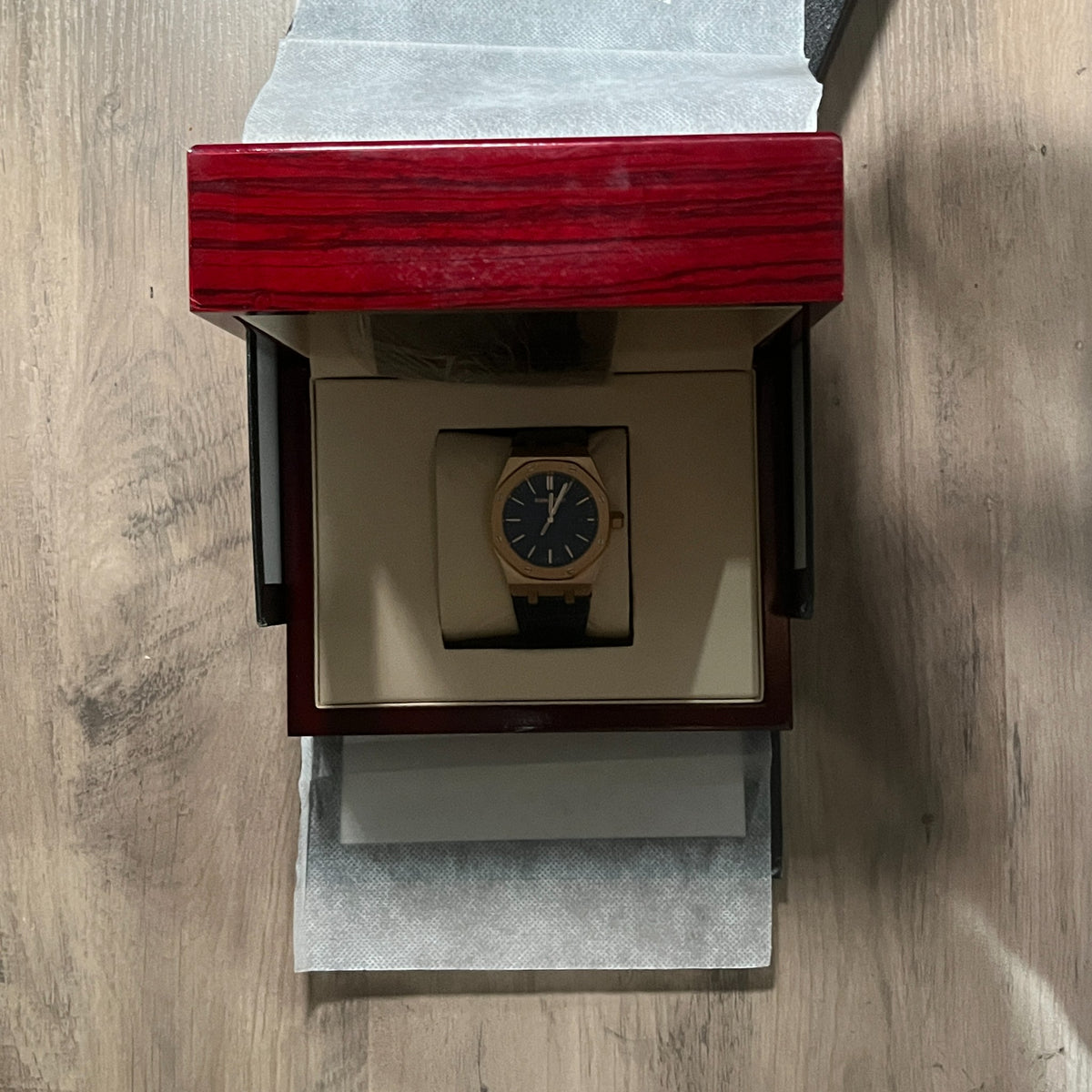 Reloj octagonal con caja en tono oro rosa, esfera azul con textura de cuadrícula y correa de piel azul, sostenido en la mano frente a una caja de presentación roja
