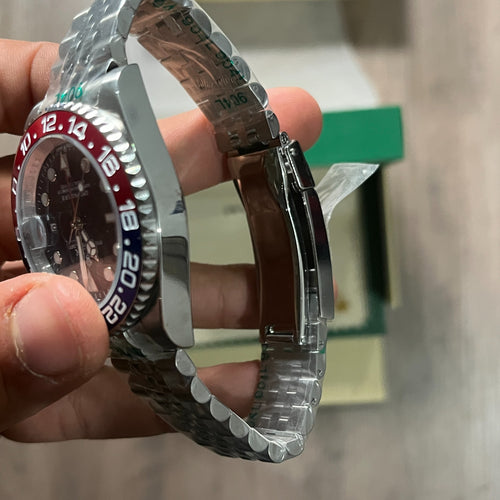 Reloj de acero con bisel azul y rojo, esfera negra con fecha y brazalete de cinco eslabones, sostenido en la mano