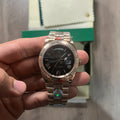 Reloj en tono oro rosa con esfera negra, bisel estriado y brazalete de eslabones, sostenido en la mano frente a una caja verde