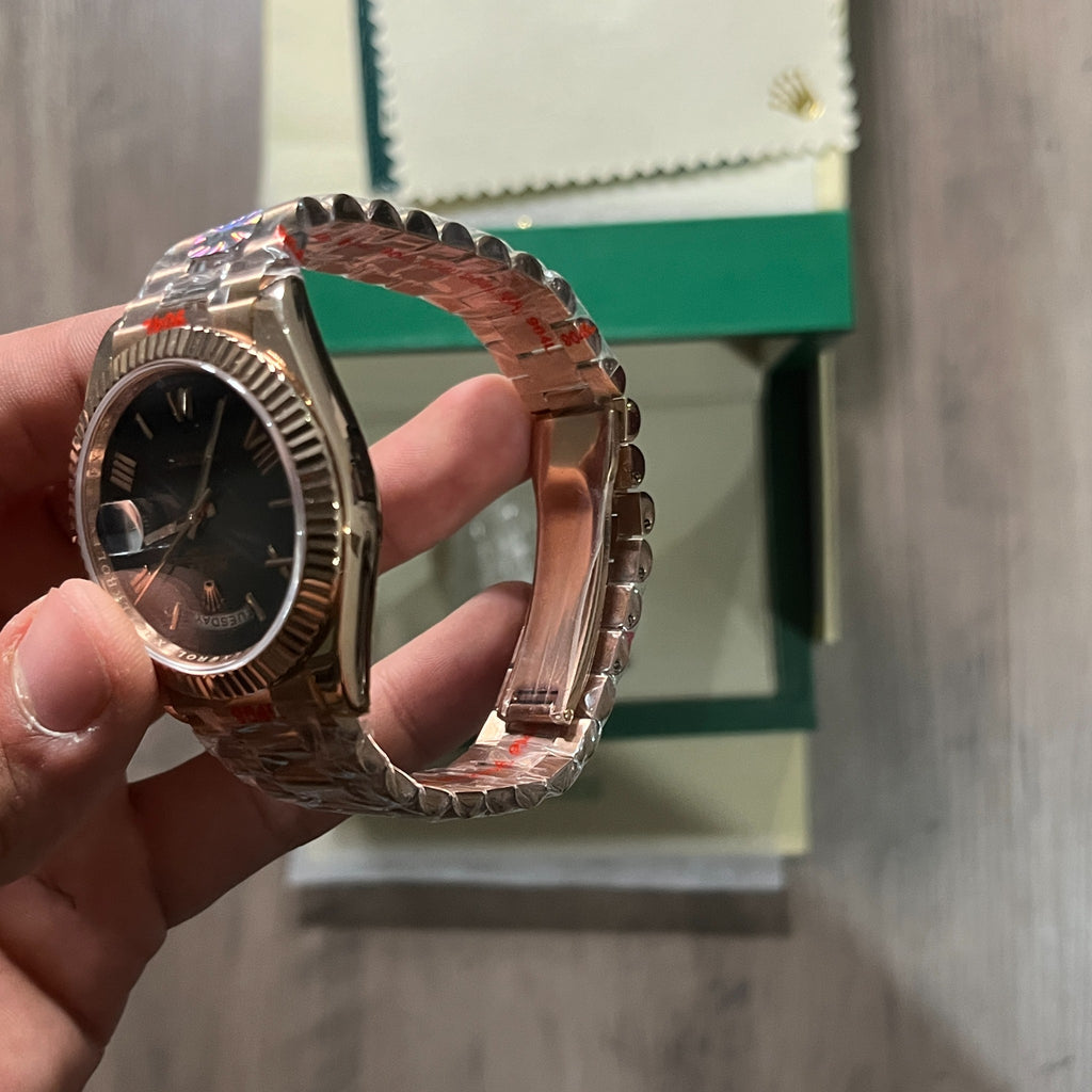 Reloj en tono oro rosa con esfera negra, bisel estriado y brazalete de eslabones, sostenido en la mano frente a una caja verde