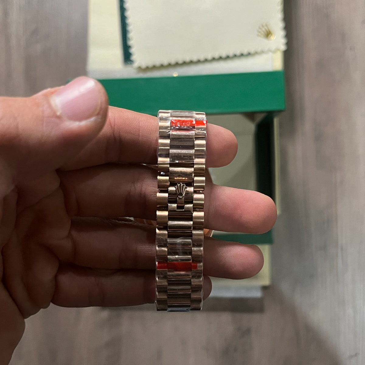 Reloj en tono oro rosa con esfera negra, bisel estriado y brazalete de eslabones, sostenido en la mano frente a una caja verde
