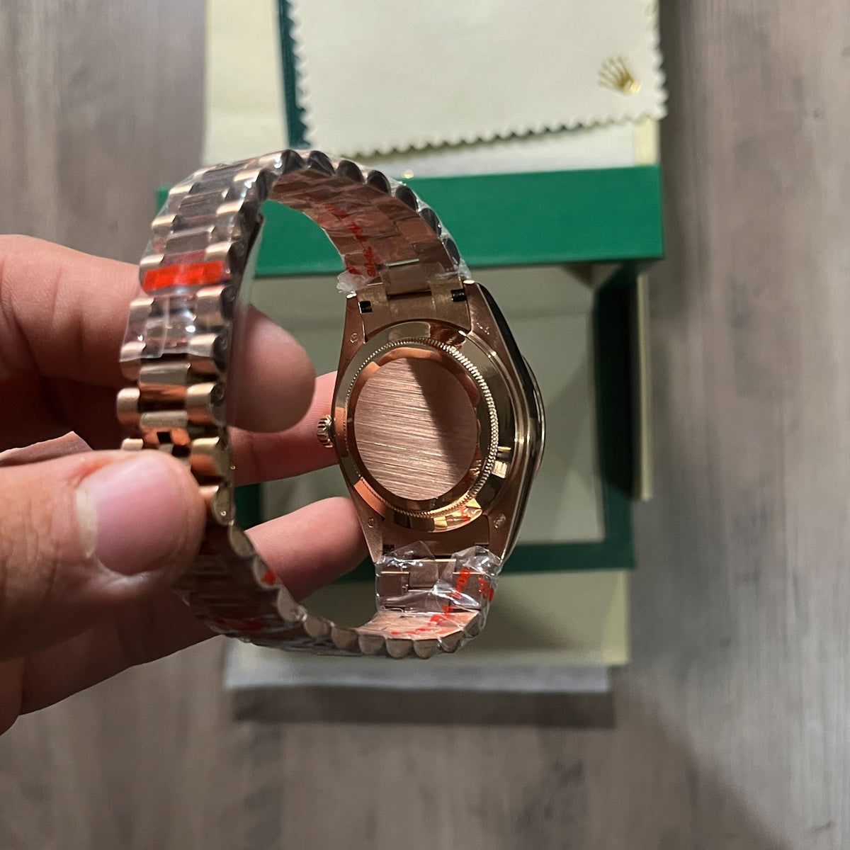 Reloj en tono oro rosa con esfera negra, bisel estriado y brazalete de eslabones, sostenido en la mano frente a una caja verde