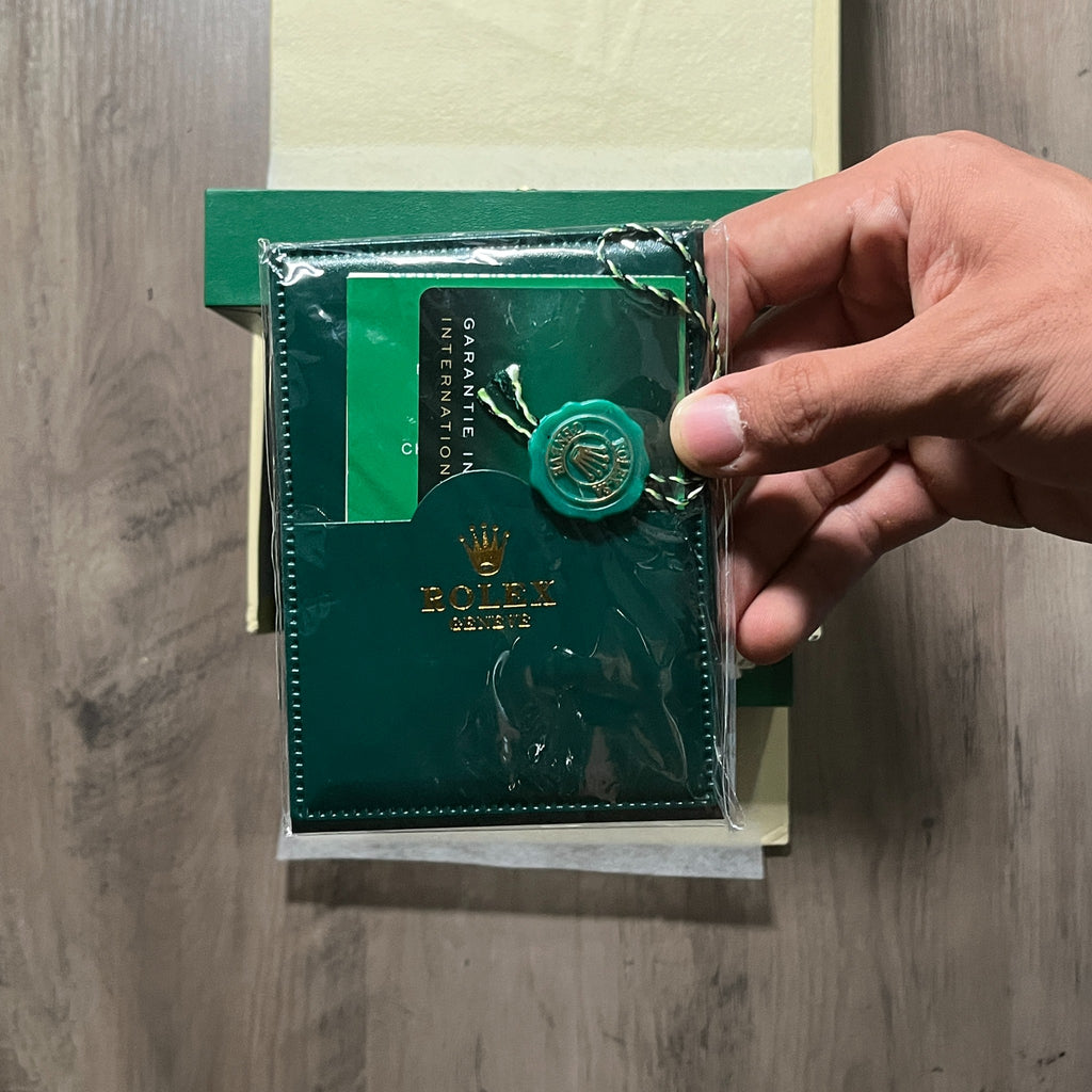 Reloj en tono oro rosa con esfera negra, bisel estriado y brazalete de eslabones, sostenido en la mano frente a una caja verde