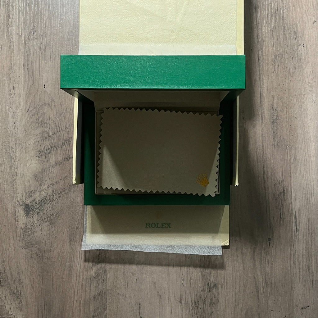 Reloj en tono oro rosa con esfera negra, bisel estriado y brazalete de eslabones, sostenido en la mano frente a una caja verde