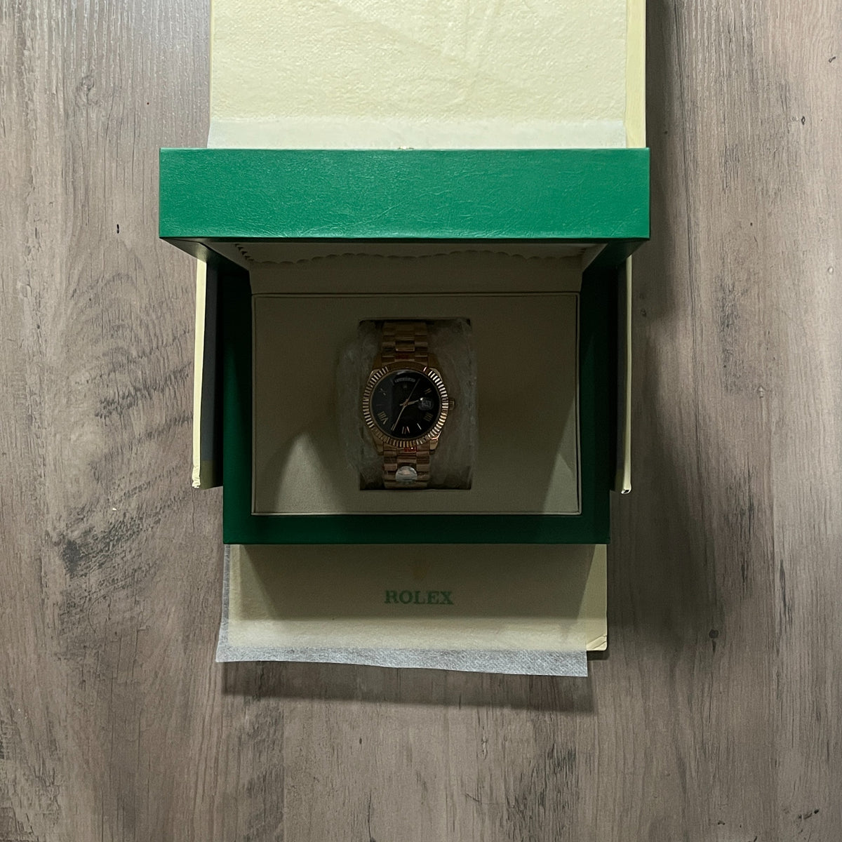 Reloj en tono oro rosa con esfera negra, bisel estriado y brazalete de eslabones, sostenido en la mano frente a una caja verde