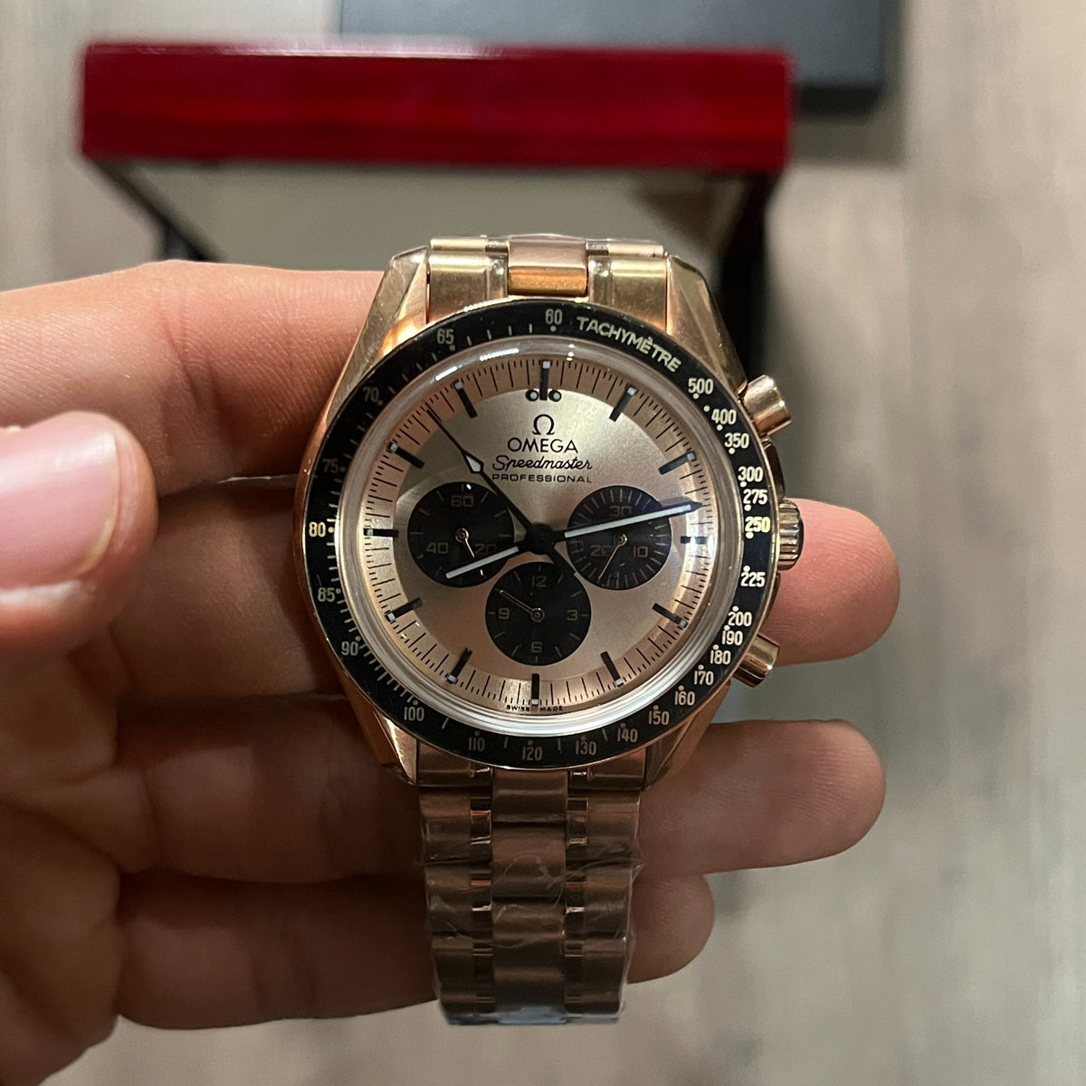 Reloj cronógrafo en tono oro rosa con bisel negro taquimétrico, esfera champagne con tres subesferas negras y brazalete de acero, sostenido en la mano frente a una caja roja