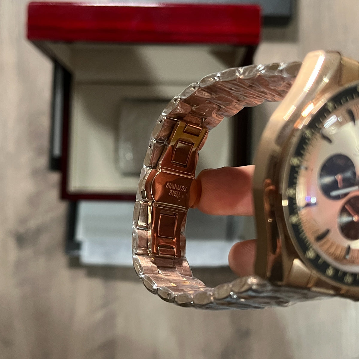 Reloj cronógrafo en tono oro rosa con bisel negro taquimétrico, esfera champagne con tres subesferas negras y brazalete de acero, sostenido en la mano frente a una caja roja