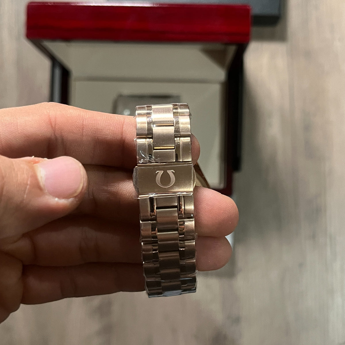 Reloj cronógrafo en tono oro rosa con bisel negro taquimétrico, esfera champagne con tres subesferas negras y brazalete de acero, sostenido en la mano frente a una caja roja