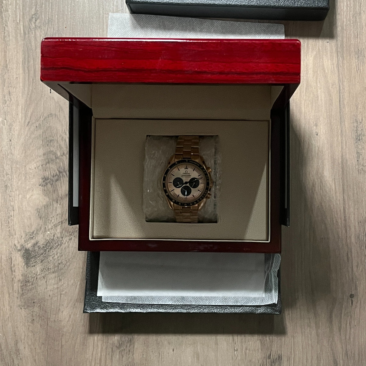Reloj cronógrafo en tono oro rosa con bisel negro taquimétrico, esfera champagne con tres subesferas negras y brazalete de acero, sostenido en la mano frente a una caja roja