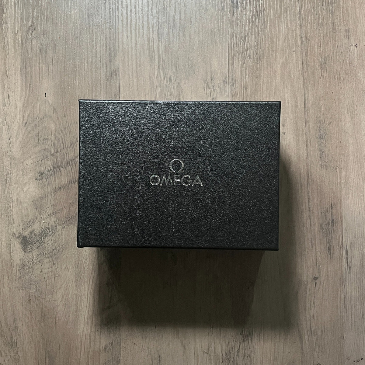 Reloj cronógrafo en tono oro rosa con bisel negro taquimétrico, esfera champagne con tres subesferas negras y brazalete de acero, sostenido en la mano frente a una caja roja