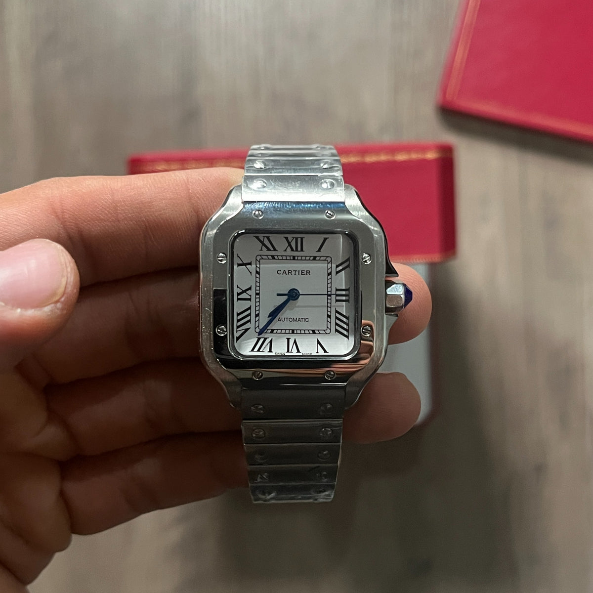 Reloj cuadrado de acero con esfera blanca, números romanos y manecillas azules, sostenido en la mano frente a una caja roja