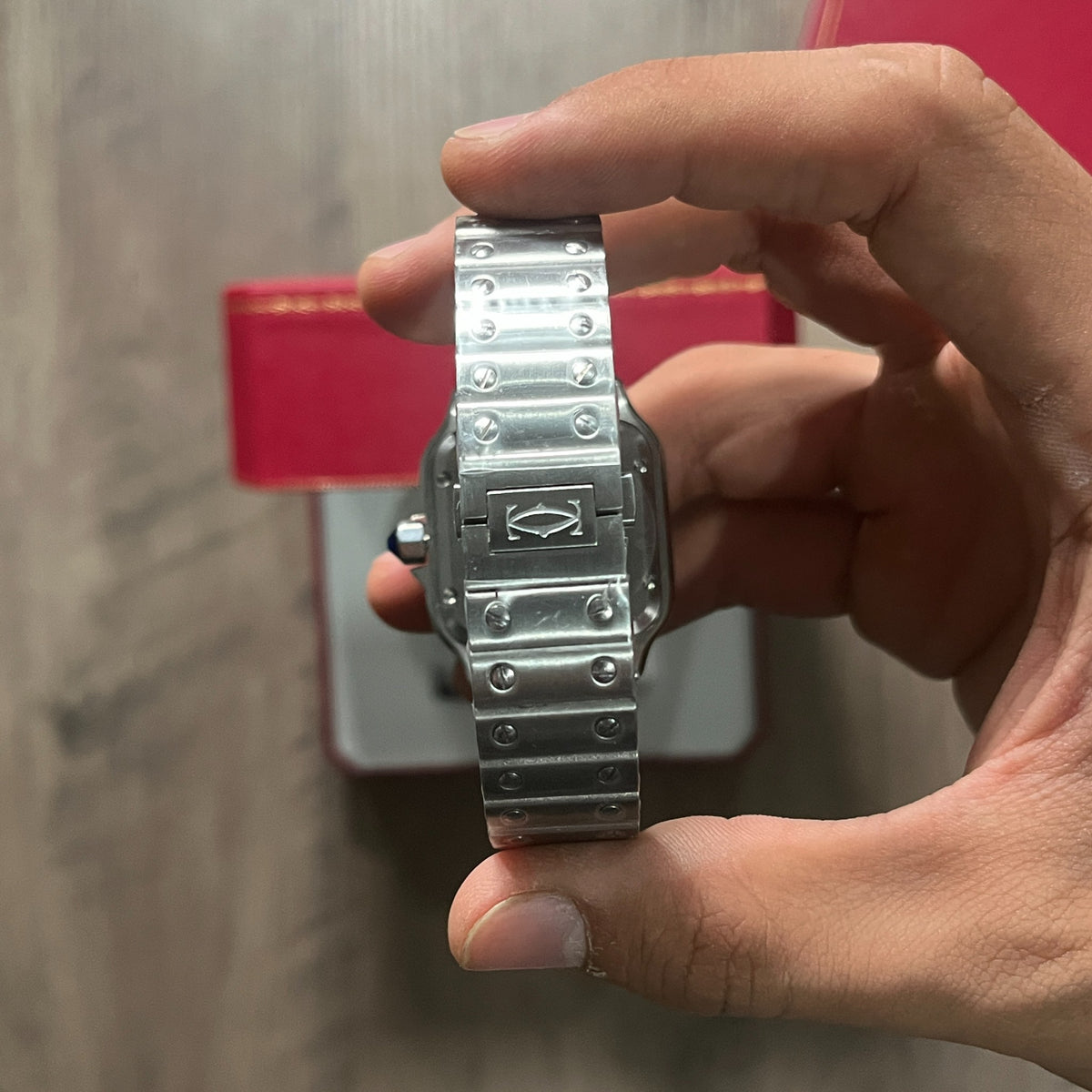Reloj cuadrado de acero con esfera blanca, números romanos y manecillas azules, sostenido en la mano frente a una caja roja