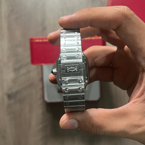 Reloj cuadrado de acero con esfera blanca, números romanos y manecillas azules, sostenido en la mano frente a una caja roja