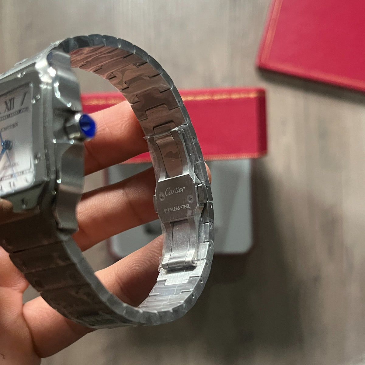 Reloj cuadrado de acero con esfera blanca, números romanos y manecillas azules, sostenido en la mano frente a una caja roja